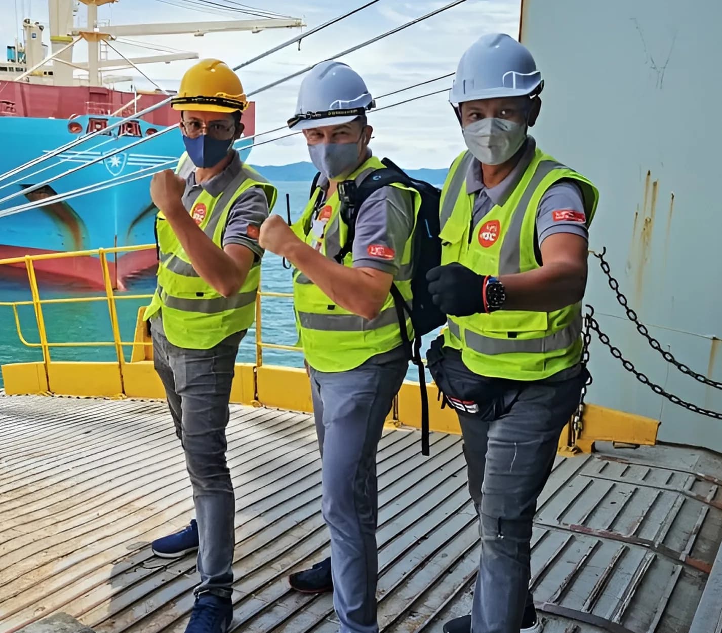 Three HSC specialists in hardhats and safety vests posing on a vessel deck beside a container ship